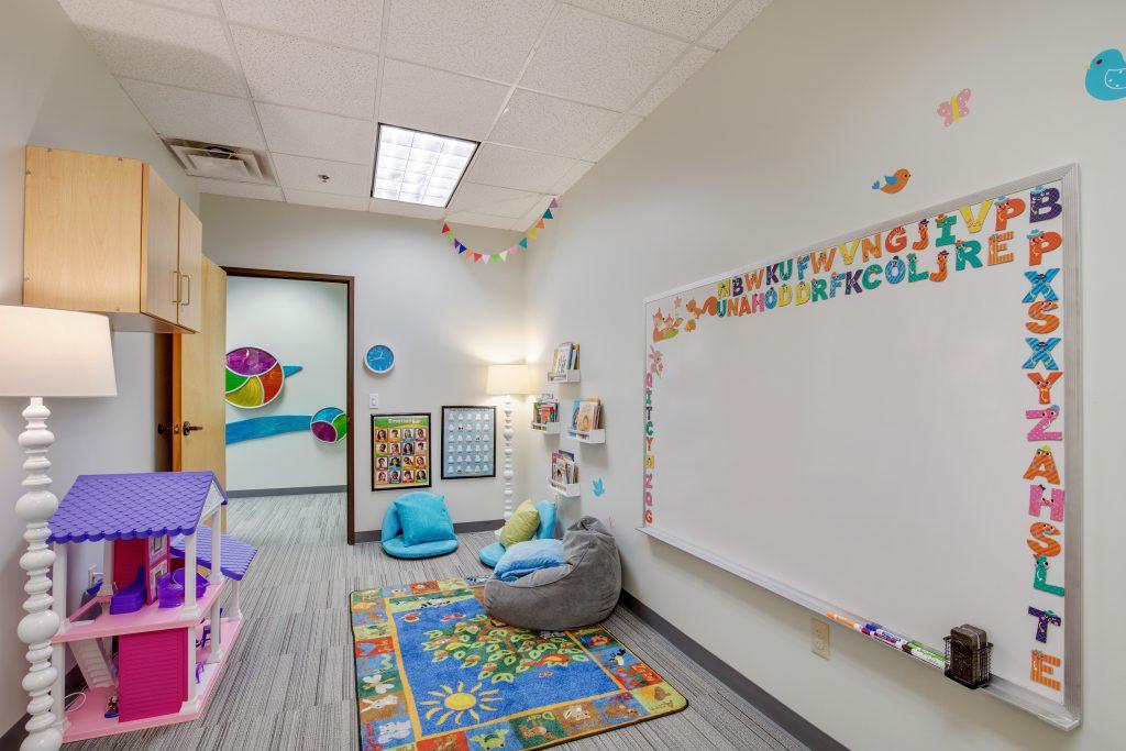 Child's playroom. Featuring doll house, white board with the alphabet, a colorful rug, and squishy seats.