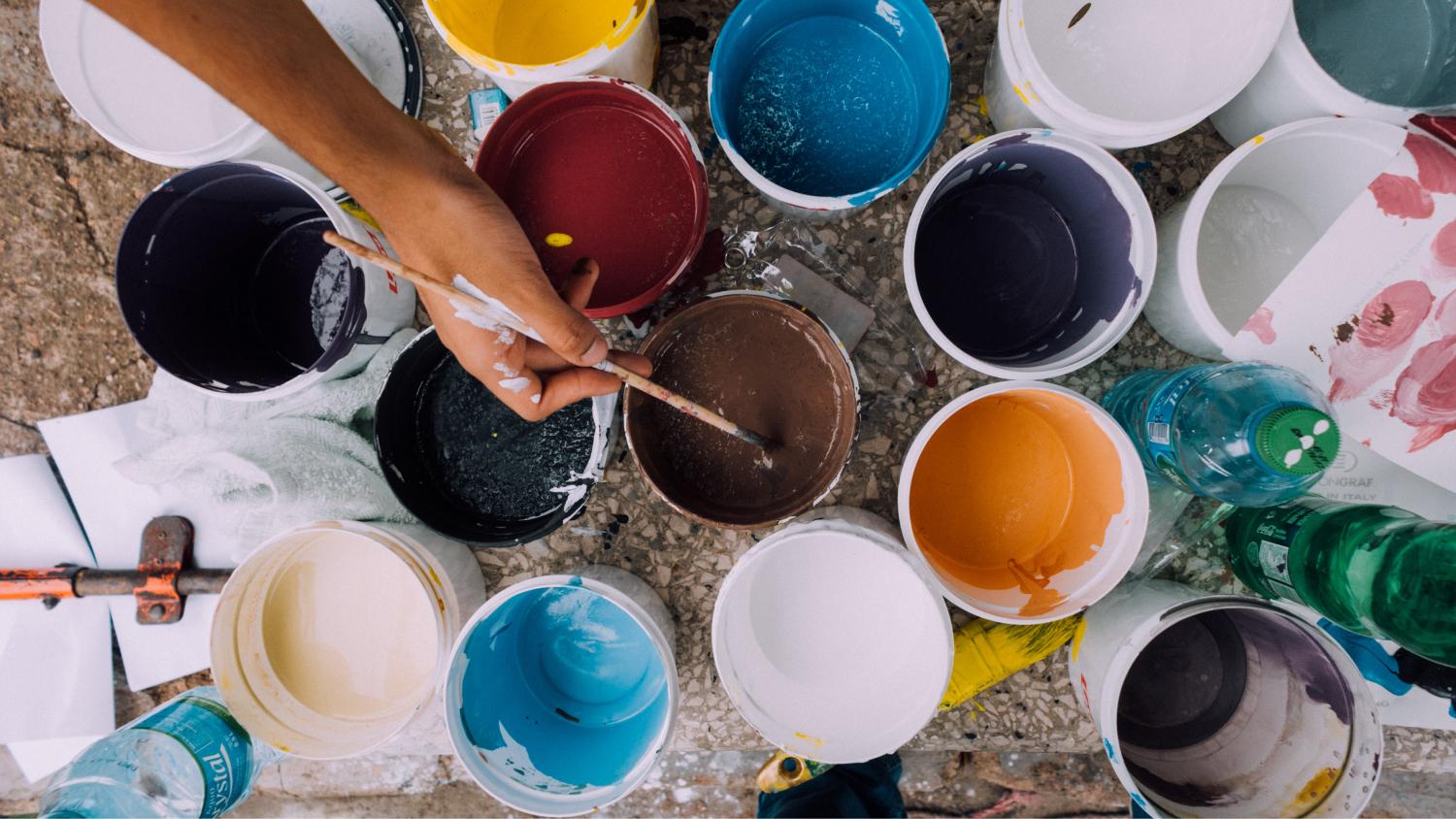 Cans of paint and a hand dipping into one with a paint brush.