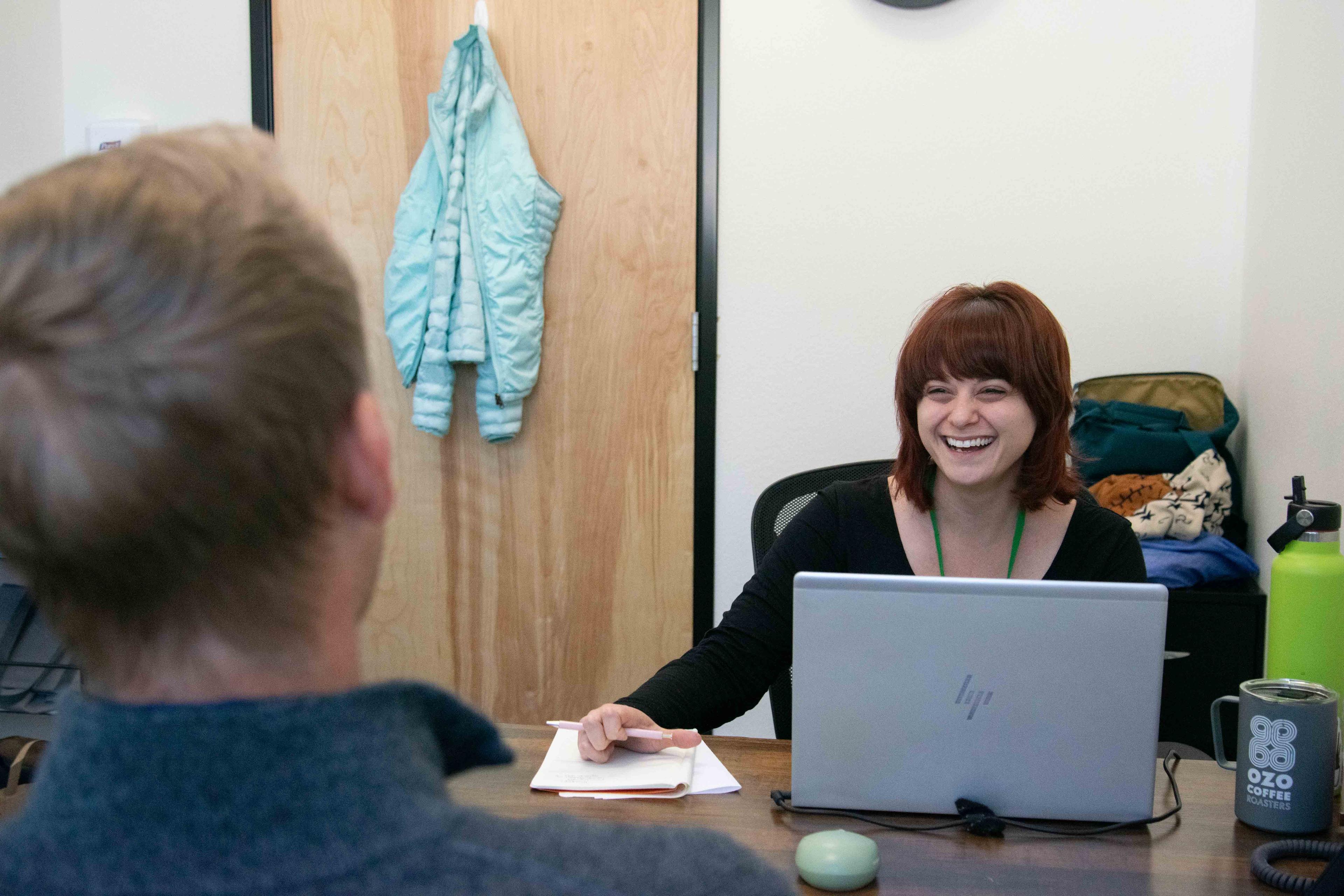 Therapist smiling and laughing sitting at desk talking to a person.