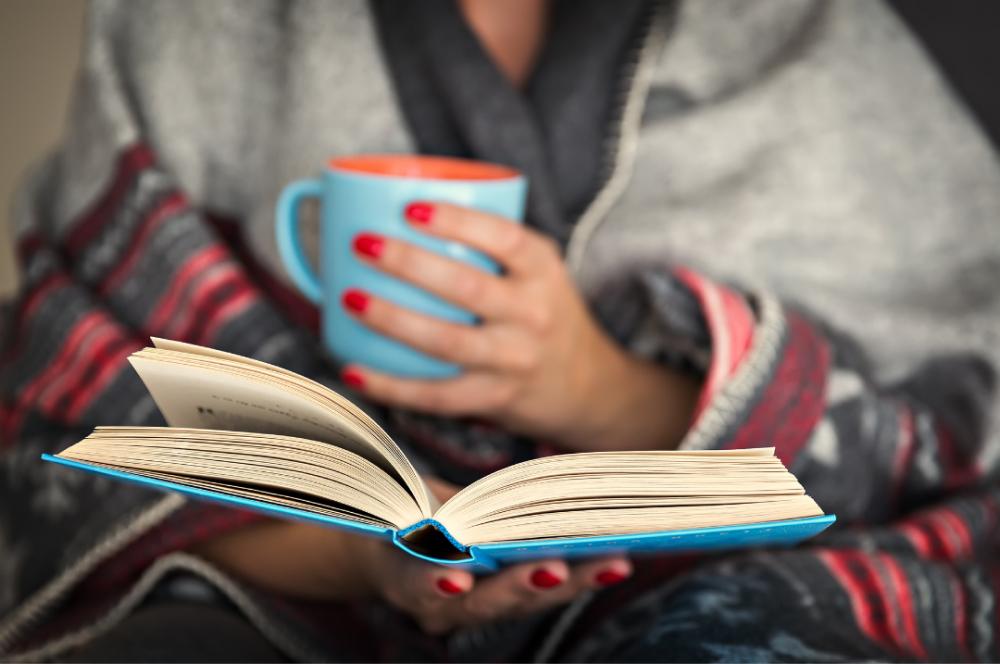 Behavioral Health Homes Tile: Person snuggled up in blanket reading book while holding a mug.