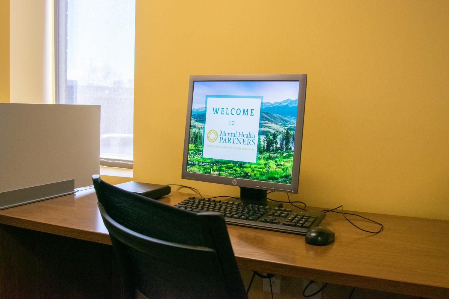 Computer lab: computer monitor on desk with chair. Text on monitor: Welcome to Mental Health Partners. 